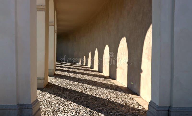 Old colonnade with the shadows of the arches on the opposite wall and cobblestone floor. stock image